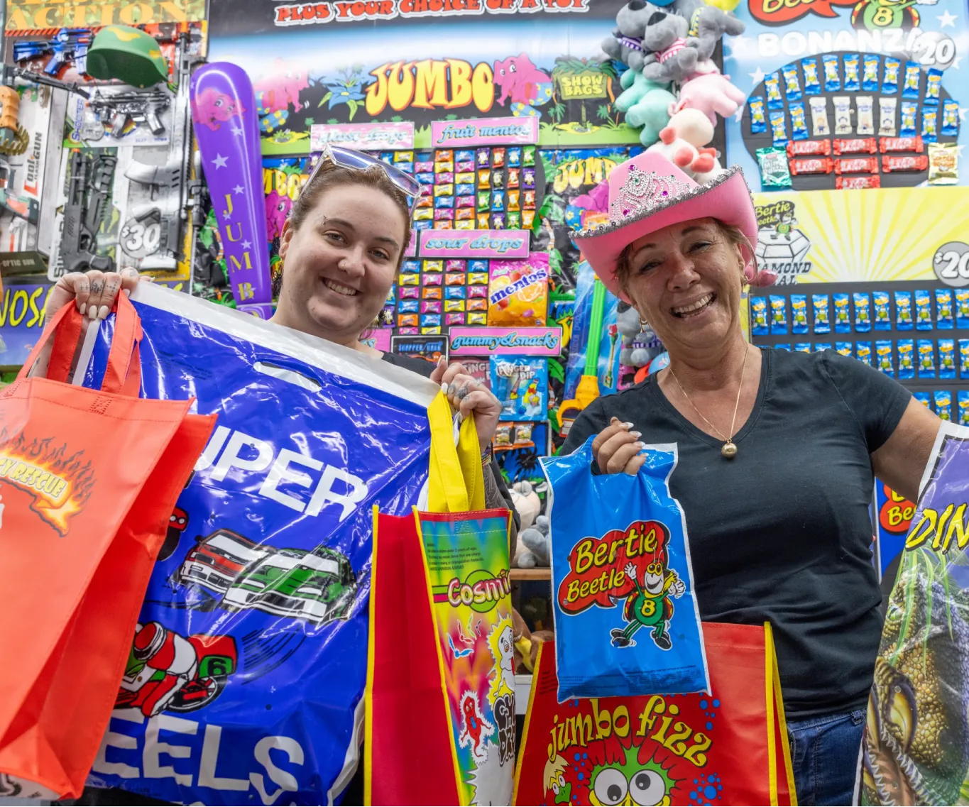 Two smiling women pose at a colorful carnival prize booth, holding oversized bags.