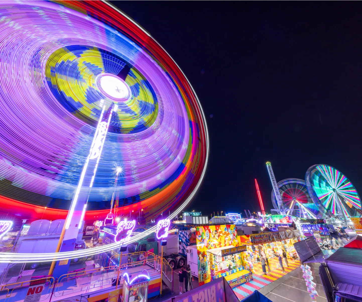 Neon-lit carnival at night, with a giant spinning ride, Ferris wheels, colorful booths, and an energetic atmosphere.